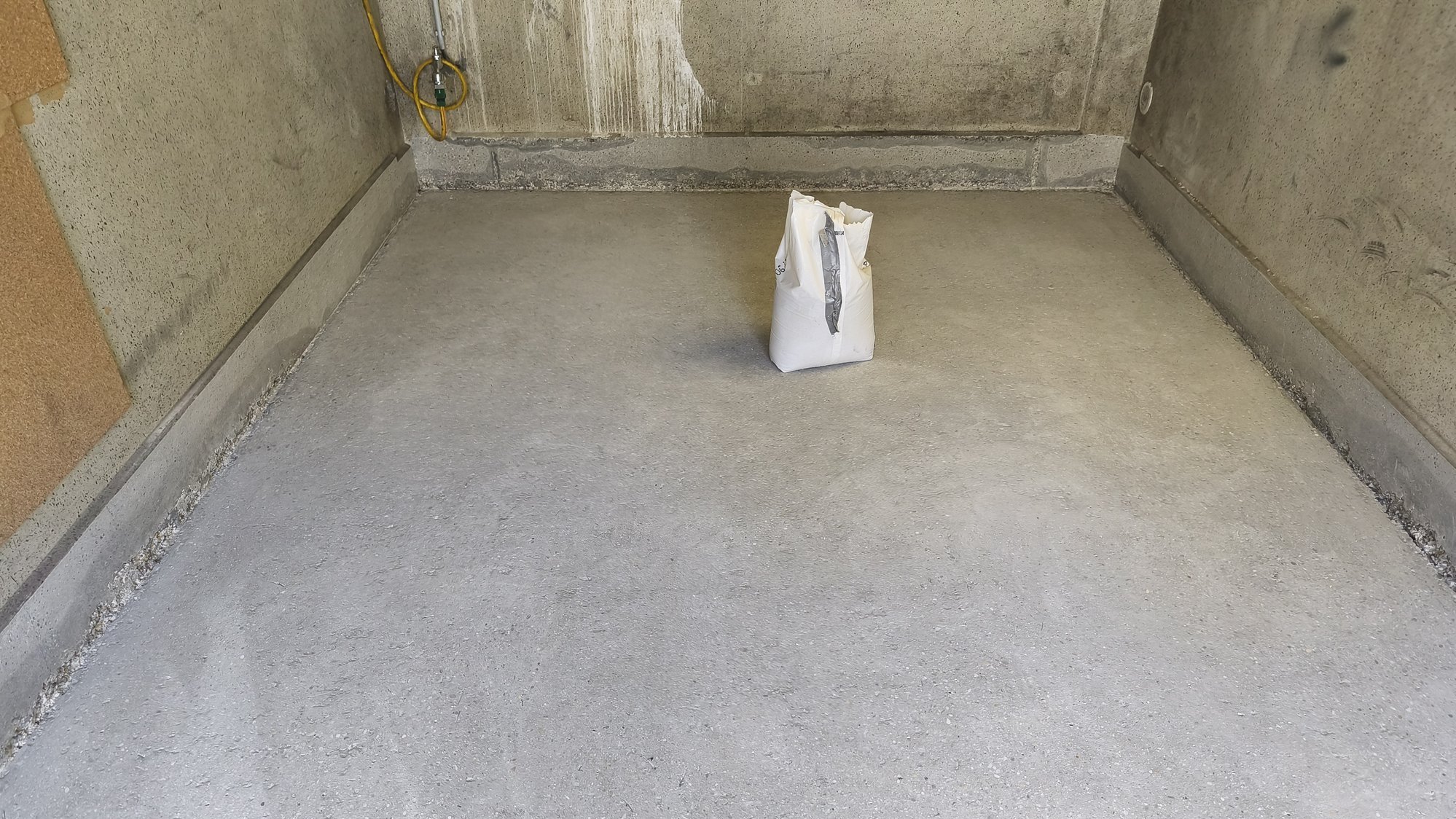 Raum mit glatt verteilter grauer Masse auf dem Boden, weißer Sack steht mittig
