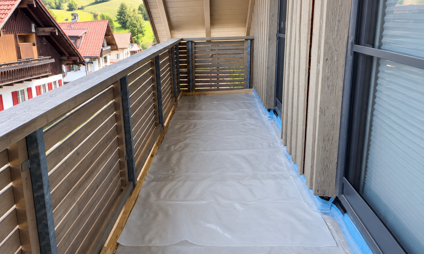 Balkon mit Holzgeländer, Boden mit transparenter Folie abgedeckt, blaues Klebeband an Fensterrahmen