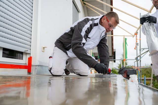 Arbeiter glättet graue Masse auf Balkonboden neben Hauswand.