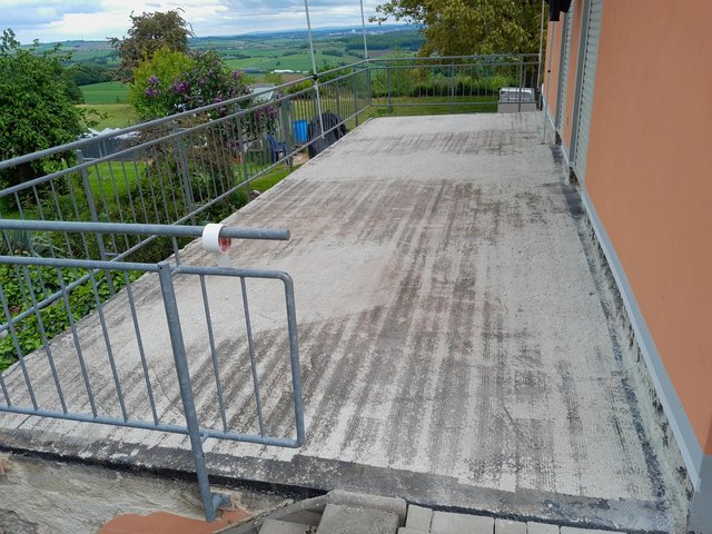 Grauer Betonbalkon mit Metallgeländer und verschmutzter Oberfläche vor Landschaftsblick