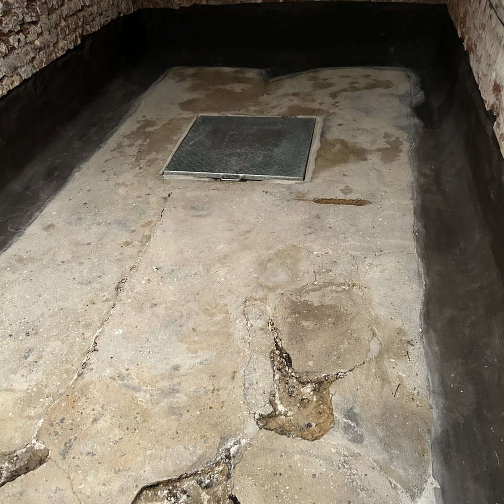 Rissiger Kellerboden aus Beton mit Metallluke, Ziegelwände mit schwarzer Masse am Sockel.