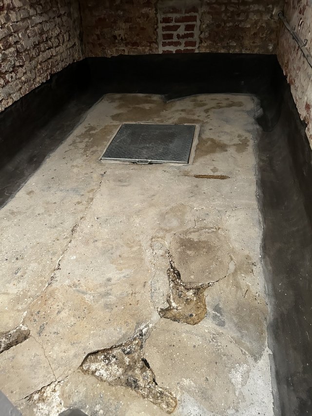 Rissiger Kellerboden aus Beton mit Metallluke, Ziegelwände mit schwarzer Masse am Sockel.
