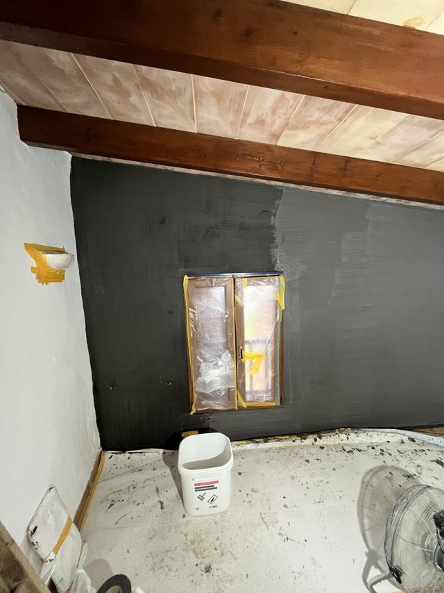 Abgeklebtes Fenster, graue Masse auf Wand aufgetragen, Eimer auf Abdeckfolie im Raum