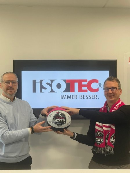 Zwei Männer halten in der Mitte gemeinsam einen signierten Basketball. Im Hintergrund das ISOTEC-Logo. 