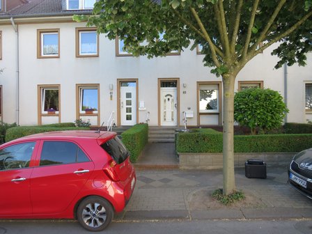 Reihenhausfassade mit zwei Haustüren, Hecken, Baum und parkendem roten Auto.
