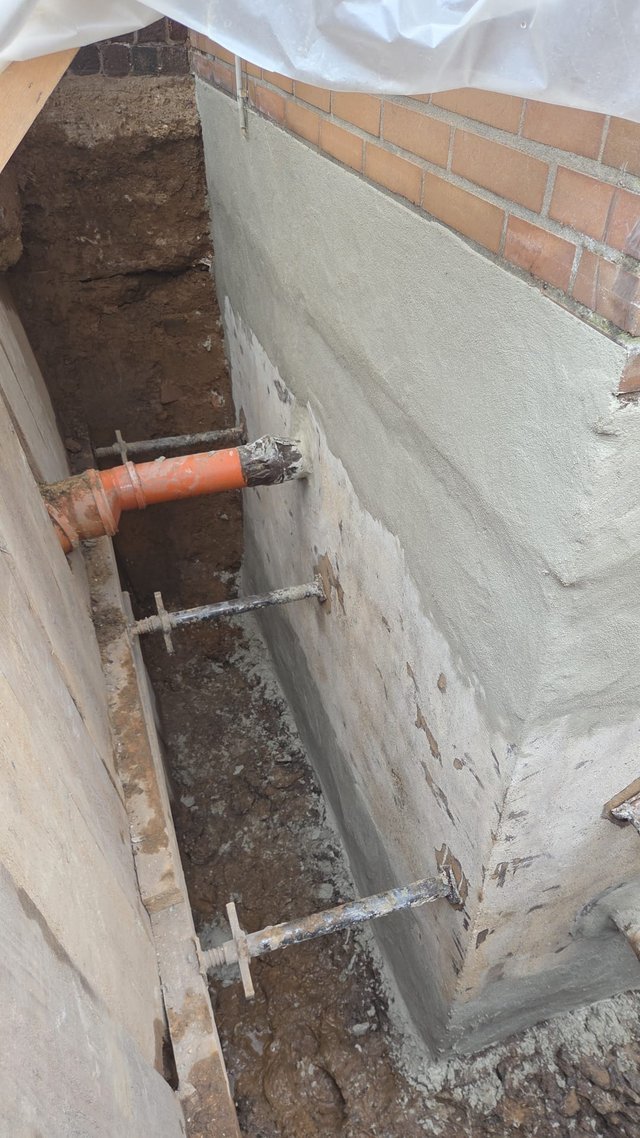 Freigelegte Kellerwand mit grauer Masse und orangefarbenem Rohr im Erdgraben