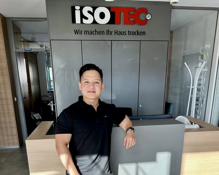 Mann steht am Empfangstresen vor ISOTEC Logo und grauen Schranktüren.