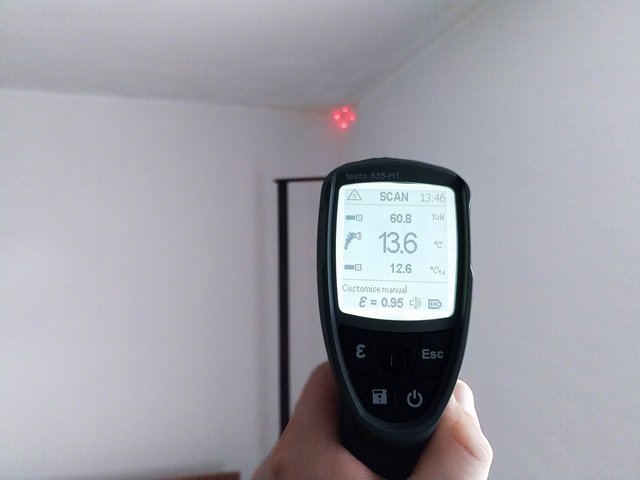 Hand hält Messgerät mit roten Laserpunkten auf weiße Zimmerecke gerichtet.