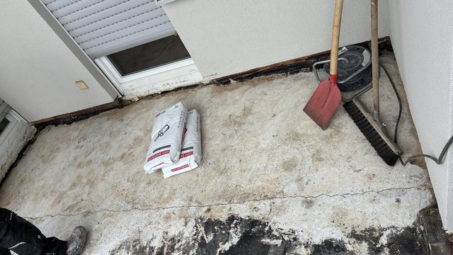 Rissiger Betonboden am Fenster, Baustoffbeutel sowie Schaufel und Besen liegen bereit