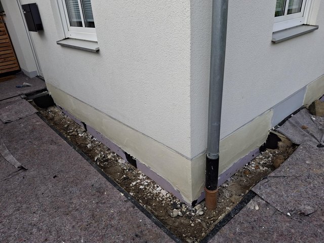 Helle Sockelabdichtung an Gebäudeecke, freigelegter Kiesstreifen am Fundament