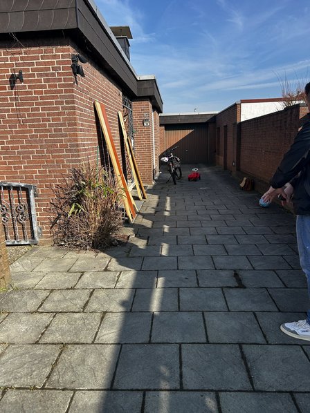 Gepflasterte Hofeinfahrt mit Backsteinwänden, Garage, Fahrrad und abgestellten Holzbrettern.