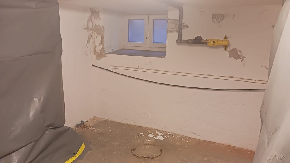 Kellerraum mit freigelegten Putzstellen, Fenster und gelber Rohrleitung; Bauschutt am Boden