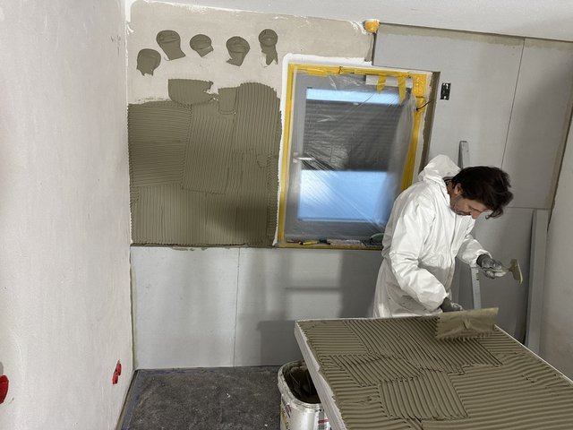 Arbeiter trägt graue Masse mit Zahnspachtel auf helle Platte auf, abgeklebt­es Fenster daneben.