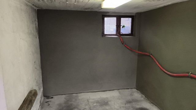 Graue Masse auf Kellerwand und Boden, roter Schlauch vor kleinem Fenster