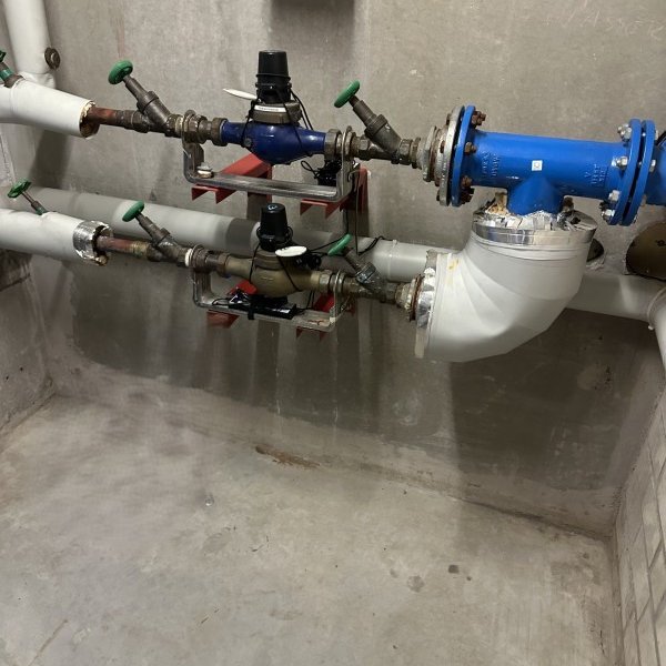 Rohrleitungen mit Armaturen und blauem Gerät an grauer Betonwand in Technikraum