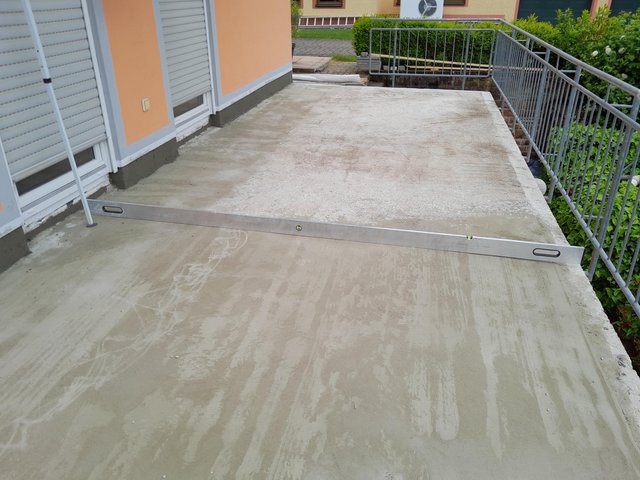 Aluminium-Wasserwaage liegt auf grauer Betonplatte am Balkon neben Metallgeländer.