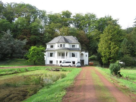 Weiße Villa am Waldrand, Zufahrt mit geparkten Autos, Teich und Grünfläche im Vordergrund
