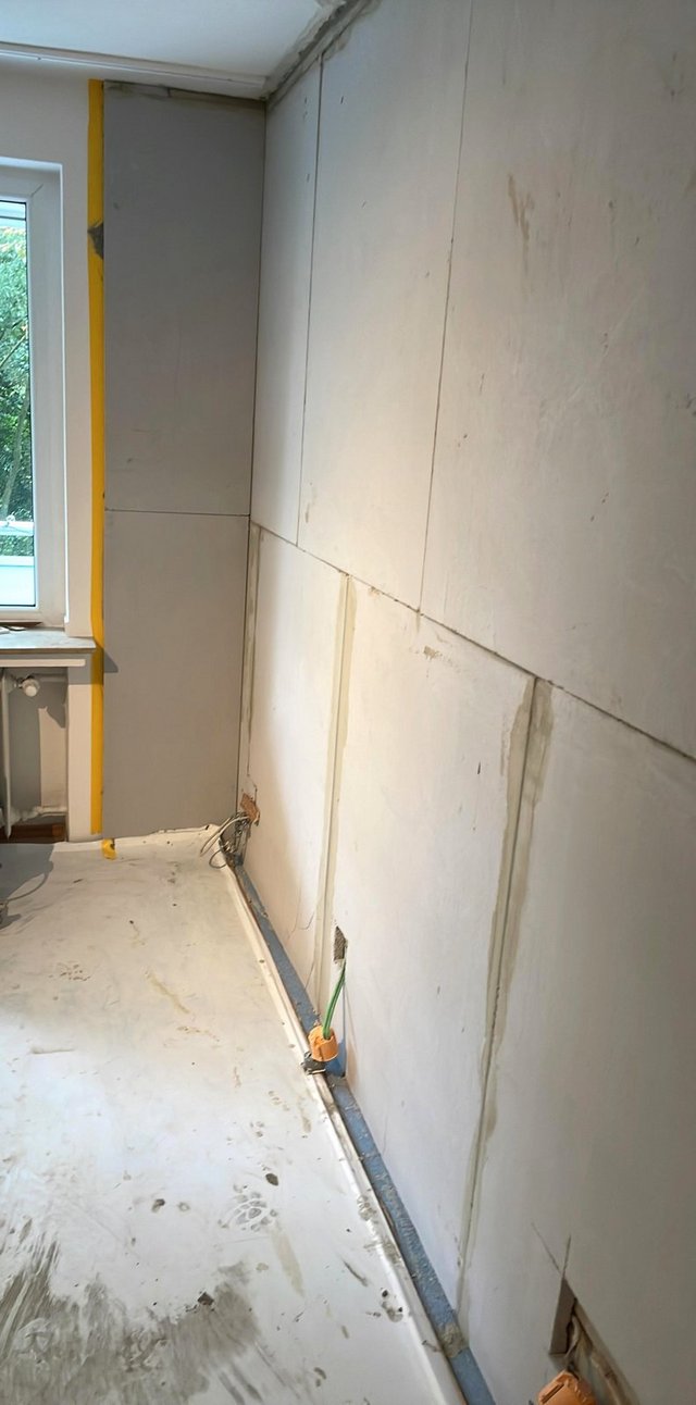 Helle Plattenverkleidung mit Fugen an Zimmerwand, sichtbare Kabel am Sockel neben Fenster.