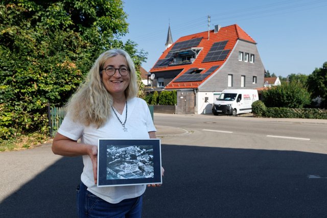 Frau hält gerahmtes Luftbild vor Haus mit Solarmodulen an Straße.