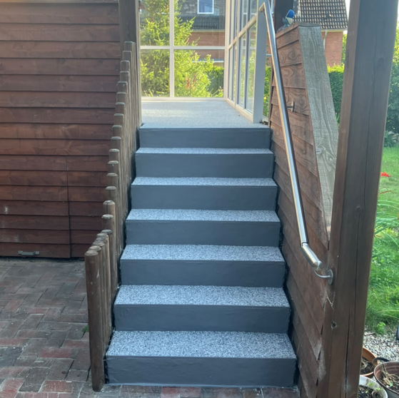 Graue Treppe mit hell gesprenkelten Stufen, Edelstahl-Handlauf und Holzverkleidung im überdachten Eingangsbereich