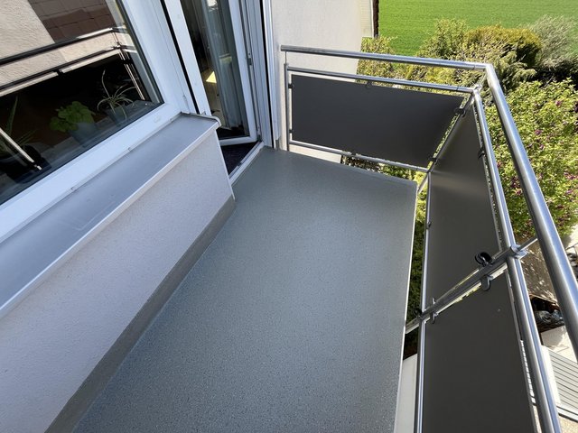 Kleiner Balkon mit grauem Bodenbelag, Metallgeländer und offener Balkontür.