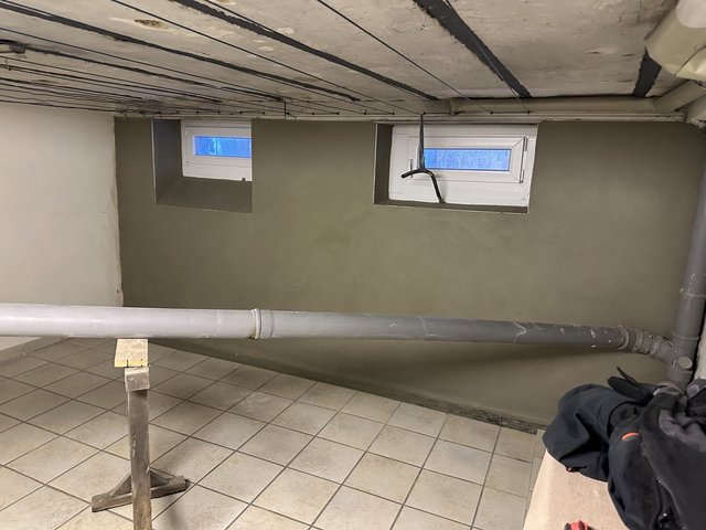 Graue Beschichtung an Kellerwand mit zwei kleinen Fenstern und freigelegten Leitungen