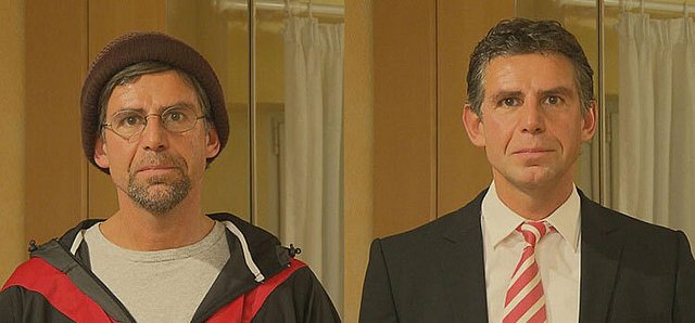 Zwei Männer stehen nebeneinander, einer sportlich, einer formell gekleidet.