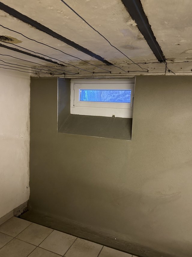 Feuchter Kellerraum mit verfärbter Decke, Kabeln und kleinem Fenster