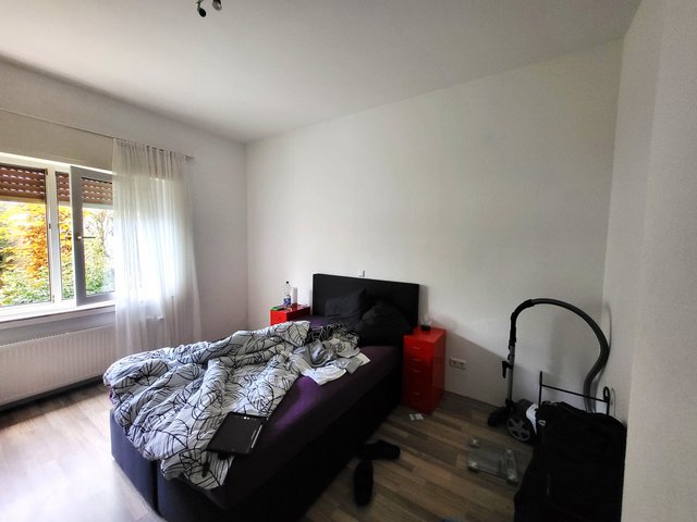 Unordentliches Schlafzimmer mit Bett, roten Nachttischen, Fenster mit Vorhang und Staubsauger