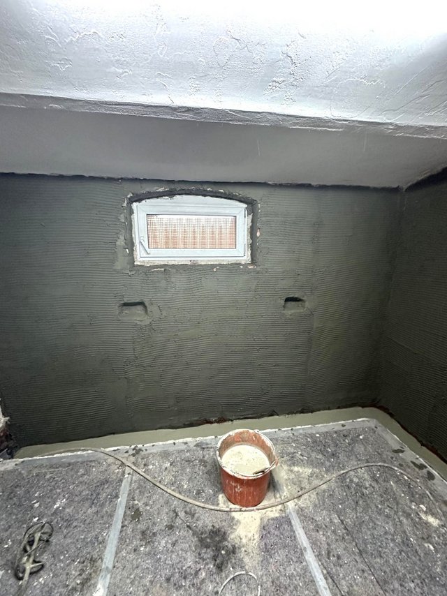 Kellerraum mit grauer Masse an Wänden, kleines Fenster und Eimer auf abgedecktem Boden