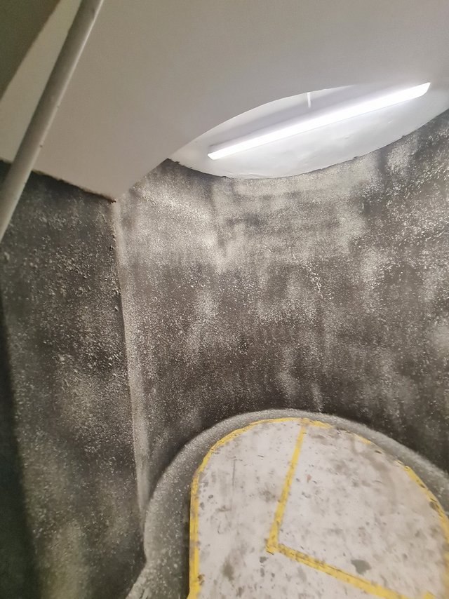Schwarzgraue Kellerwand mit weißen Flecken, daneben runder Betondeckel mit gelber Markierung