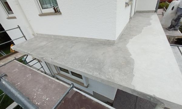 Grauer Balkonboden mit abgegrenzter, dunklerer Fläche neben weißer Fassade.
