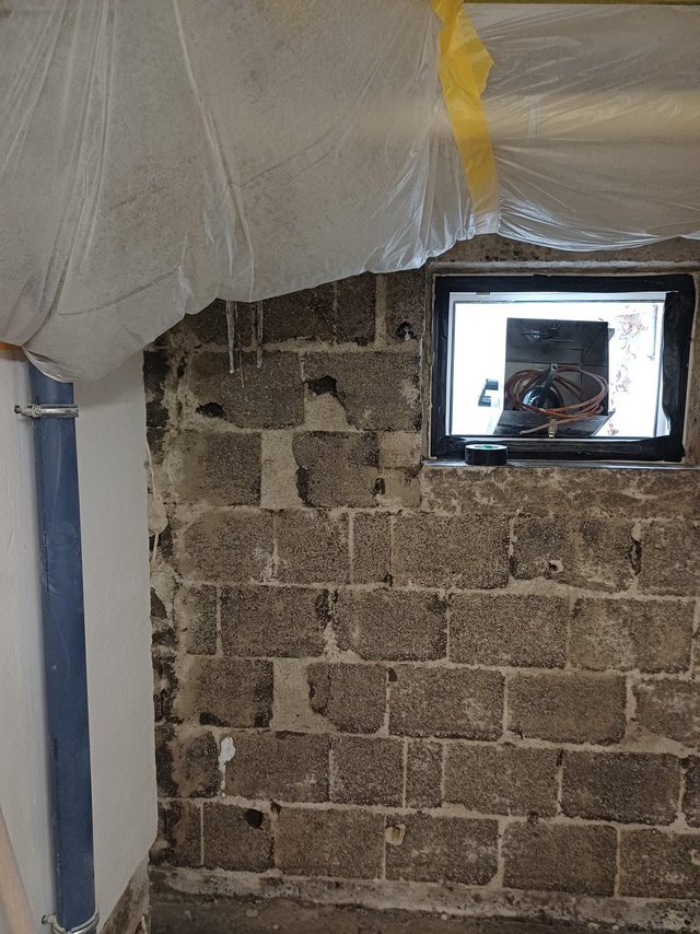 Freigelegte Kellerwand aus grauen Betonsteinen mit kleinem Fenster und Folienabdeckung.