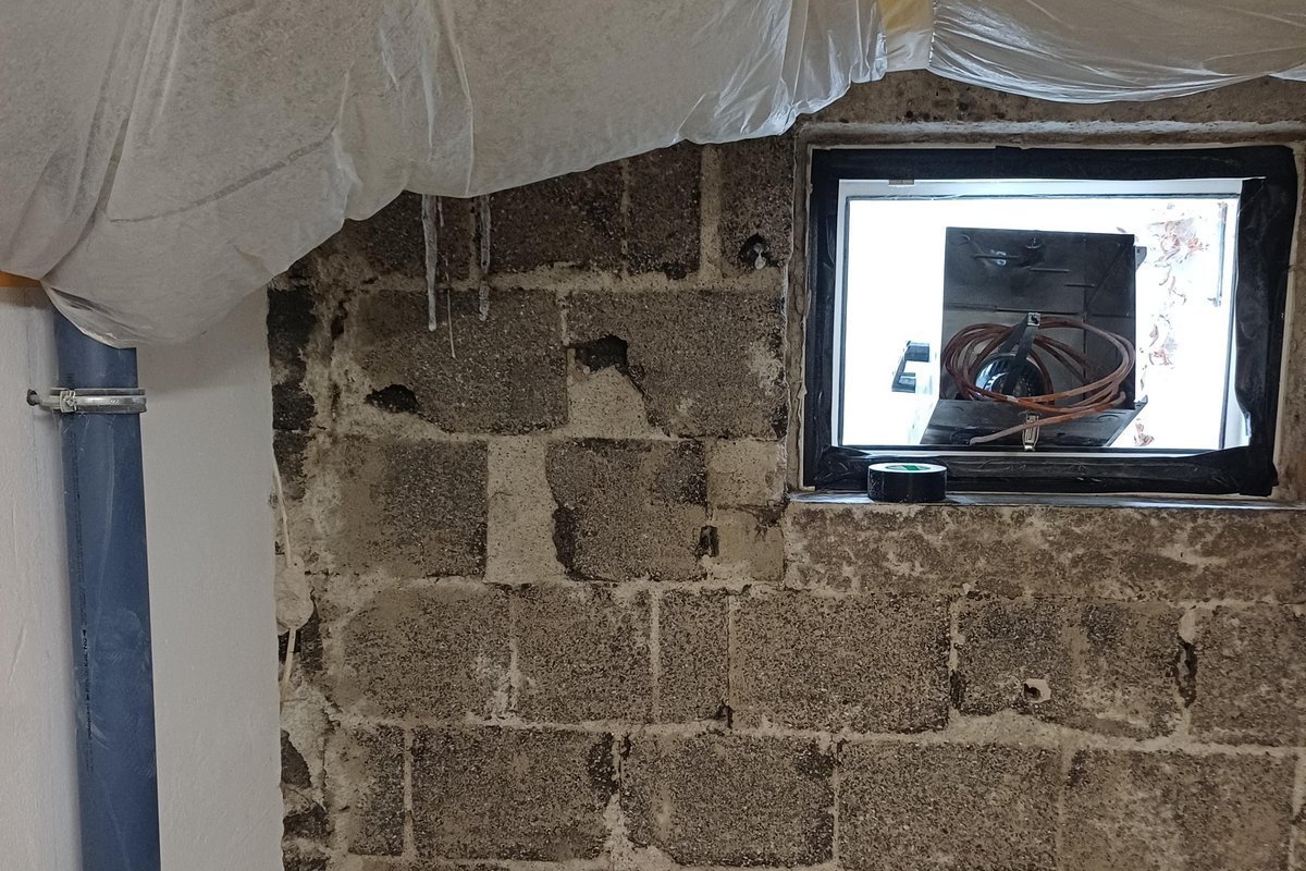 Freigelegte Kellerwand aus grauen Betonsteinen mit kleinem Fenster und Folienabdeckung.