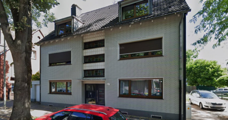 Mehrfamilienhaus mit weißer Klinkerfassade, Dachgauben und Rollläden an der Straße