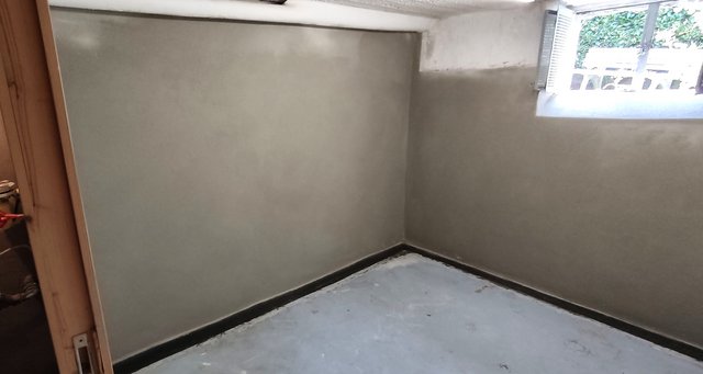 Leerer Kellerraum mit grauer Wandbeschichtung, Ecke und kleinem Fenster mit Licht.