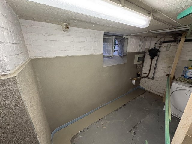 Kellerraum mit gestrichener Ziegelwand, Lüftungsfenster, Leitungen und Waschmaschine.