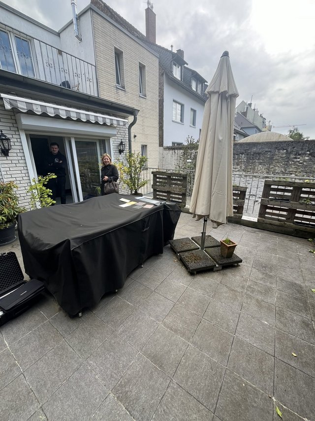 Zwei Personen stehen auf einer gefliesten Terrasse vor geoeffneter Schiebetuer und Tisch mit schwarzer Abdeckung.