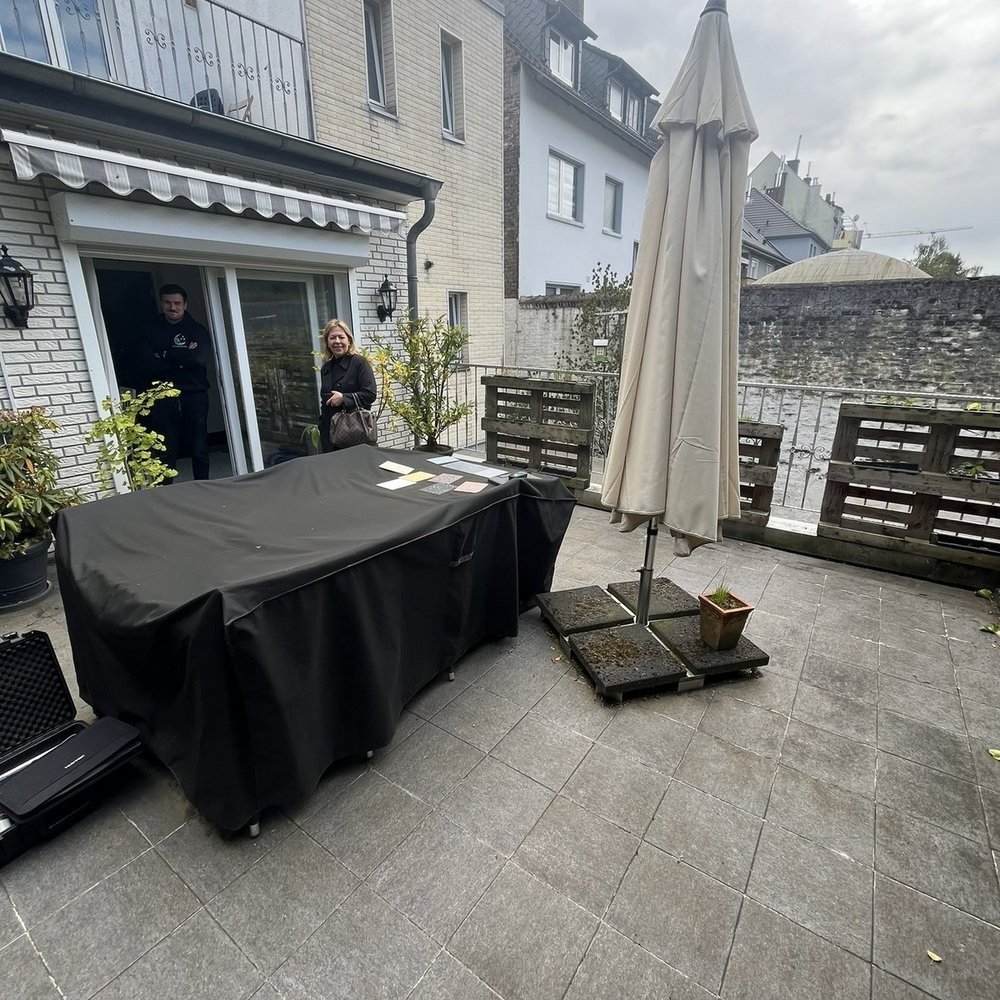 Zwei Personen stehen auf einer gefliesten Terrasse vor geoeffneter Schiebetuer und Tisch mit schwarzer Abdeckung.