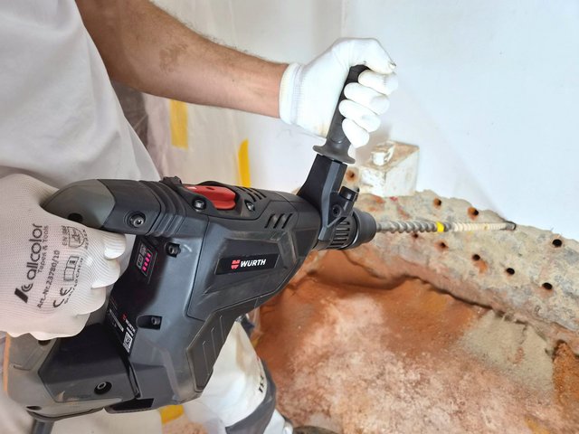 Handschuhe halten Bohrhammer mit langem Bohrer vor Betonfläche mit Bohrlöchern.