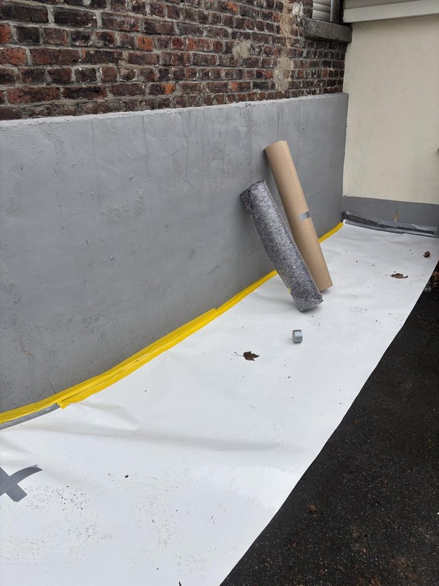 Weiße Folie mit gelbem Klebeband an grauer Mauer, Rollen lehnen an Ziegelwand.