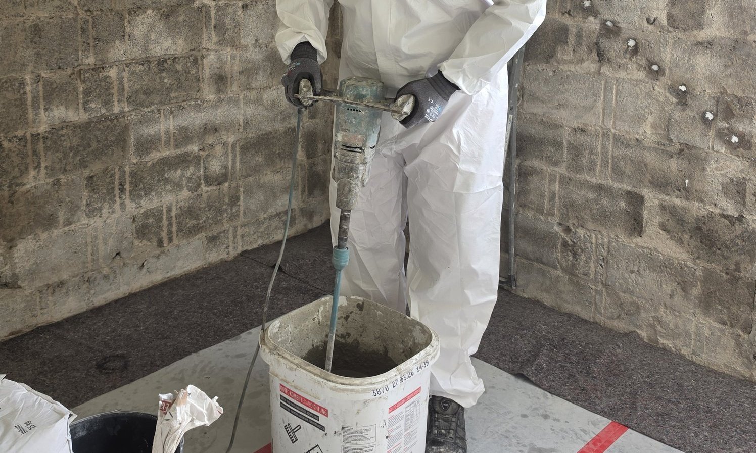 Arbeiter mischt graue Masse mit Rührwerk in Eimer vor Betonsteinwand.