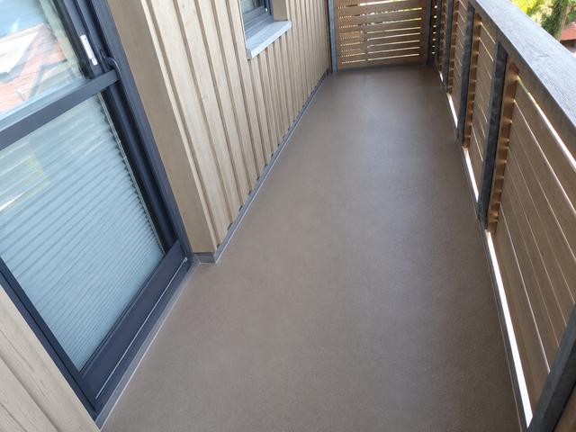 Leerer Balkon mit braun-grauem, strukturiertem Bodenbelag neben heller Holzfassade und Lamellengeländer