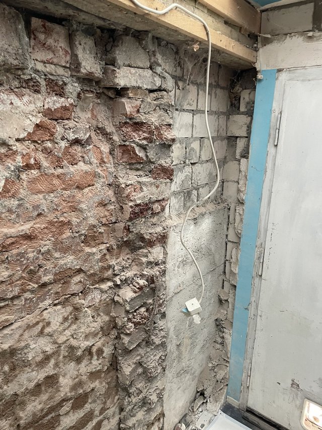 Freigelegte Ziegelwand mit bröckelnder grauer Masse, weißes Kabel führt zu einer Abzweigdose an der Wand.
