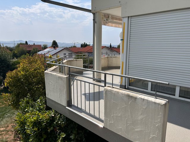 Balkon mit grauem Boden, Metallgeländer und Rollladen, Blick über Wohnhäuser und Hügel