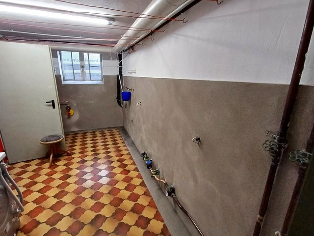 Kellerraum mit rot-gelb gemusterten Fliesen, grauer Wand und Rohrleitungen mit Wasserzähler