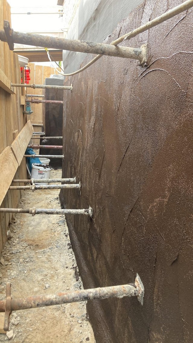 Schwarze Beschichtung auf Betonwand in Baugrube, mit Geruestankern und Kiesboden