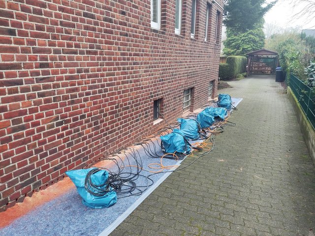 Bohrlöcher in Ziegelwand, Kabel und blaue Säcke entlang gepflastertem Hauszugang