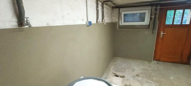 Glatte graue Beschichtung an Kellerwand mit Rohrleitungen, Fenster und Holztuer.