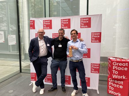 Drei Männer posieren vor „Great Place To Work“-Fotowand, einer hält einen Award.